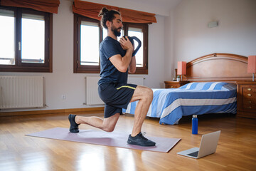Bearded man doing kettlebell lunges to work out at home while watching an online class on the laptop.