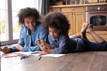 Loving African American mom and small teenage daughter lying on floor in kitchen at home paint draw together. Caring happy mother and teen girl child involved in artistic activity. Hobby concept.