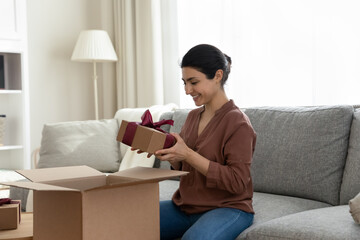 Overjoyed millennial Indian woman unpack box with wrapped birthday present for celebration. Smiling young ethnic female open unbox parcel with surprise package, excited with gift delivery.