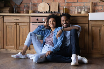 Wall Mural - Happy millennial African American couple sit on wooden floor in cozy renovated kitchen moving in together. Smiling young ethnic man and woman spouses relax at home relocate to own house. Rent concept.