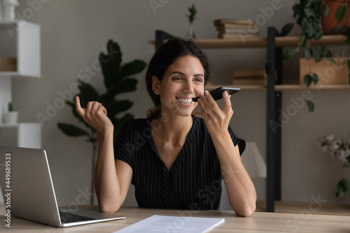 Smiling businesswoman recording audio voice message on smartphone, sitting at desk with laptop, happy woman holding phone near mouth, speaking, activating digital assistant, searching in internet