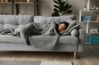 © fizkes - Full length peaceful woman resting under warm blanket on cozy couch in living room, calm young female with closed eyes sleeping, taking nap or daydreaming, lying on soft pillow with hands under cheek