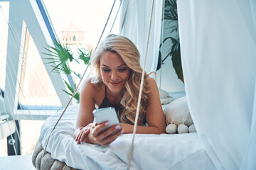 Wall Mural - Beautiful young woman using smart phone and smiling while relaxing on bed at home