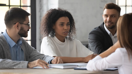 Young multiracial employees sit at desk in office engaged in team discussion, talk brainstorm together. Diverse multiethnic businesspeople discuss company business ideas at meeting. Teamwork concept.