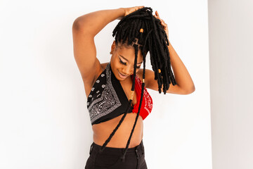 Wall Mural - Positive modern black woman in fashion swag clothes straightens dreadlocks hairstyle. Smiling african girl in trendy top bandana in vintage stylish jeans posing on white background indoors.