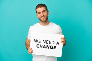 Wall Mural - Handsome blonde man over isolated blue background holding a placard with text We Need a Change with happy expression