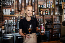 Wine Tasting. Front View Of A Female Wine Connoisseur In A Modern Uniform Working Behind The Bar. Blonde Woman Holds A Bottle Of Wine In One Hand And A Glass Full Of White Wine In The Other