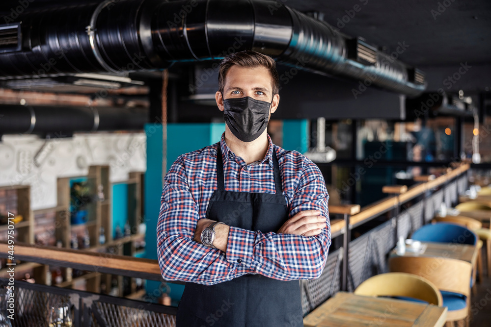 Welcome to the restaurant at the time of the corona. A male waiter with face mask stands in a cafe and wears a plaid shirt and apron. He has his arms crossed and is looking straight at the camera - obrazy, fototapety, plakaty 