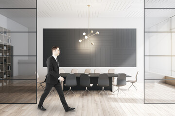 Poster - Businessman walking in modern meeting room interior with large table, wooden flooring, equipment, glass windows and sunlight.