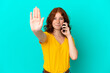 © luismolinero - Teenager reddish woman using mobile phone isolated on blue background making stop gesture