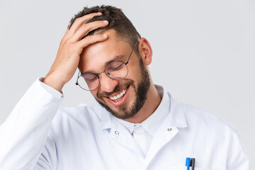 Wall Mural - Healthcare workers, coronavirus, covid-19 pandemic and insurance concept. Headshot of handsome young doctor in white coat and glasses, touching hair and laughing, smiling pleased