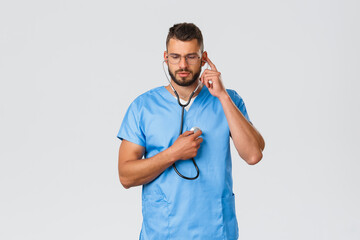 Wall Mural - Healthcare workers, medicine, covid-19 and pandemic self-quarantine concept. Serious determined doctor in blue scrubs, physician during daily checkup of patient, using stethoscope