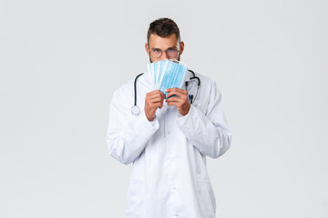 Wall Mural - Healthcare workers, medical insurance, pandemic and covid-19 concept. Funny handsome doctor, hispanic physician in white coat and glasses, showing medical masks, use it during coronavirus