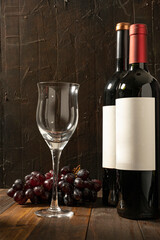 Empty wine glass next to two bottles of red wine on a rustic wooden table and dark background.