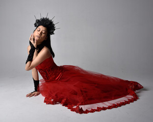 Poster - Full length  portrait of beautiful young asian woman wearing red corset and ornate gothic queen crown. Graceful sitting posing  isolated on studio background.