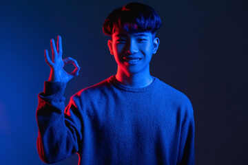 Wall Mural - Okay gesture. Approval sign. Perfect choice. Cyberpunk style. Happy asian man guarantees best quality with OK hand symbol in red blue color neon light on dark gradient background.