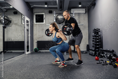 Barbell squats with the help of personal training. Fit woman in sportswear and in good shape does barbell squats to strengthen the muscles of the whole body. Coach assistance in individual training