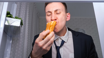 Wall Mural - Hungry overworked businessman eats cucumber with cakes and takes plate of snacks in dark kitchen at night close view from inside refrigerator