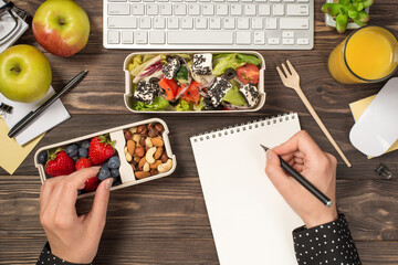 First person top view photo of hands taking blueberry from lunchbox healthy food and writing in notepad apples glass of juice plant stationery keyboard mouse isolated wooden background with copyspace