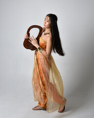 Full length portrait of pretty young asian woman wearing golden Arabian robes like a genie, holding a small musical harp, isolated on studio background.