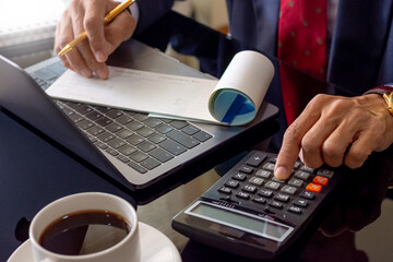 Businessman or manager in black suit with necktie hand using calculator to calculate business income,plan to paycheck , work on laptop computer and cup of coffee on the desk at modern office.