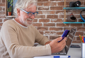 Portrait of smiling senior man using mobile phone and laptop computer at home . Brick wall on background