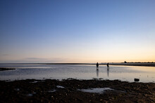 Silhouettes Of Two People Wading In Shallow Waters During Low Tide At The North Sea At Sunset