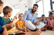 © Halfpoint - Group of small nursery school children with man teacher sitting on floor indoors in classroom, playing.