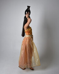 Full length portrait of pretty young asian woman wearing golden Arabian robes like a genie, standing pose holding a vase, isolated on studio background.