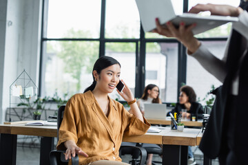 asian businesswoman talking on mobile phone near blurred colleague with laptop