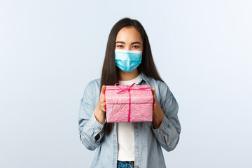 Social distancing lifestyle, covid-19 pandemic, celebrating holidays during coronavirus concept. Cute asian girl in medical mask prepared present for friends birthday, give pink gift box