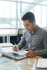 Wall Mural - Serious young UI designer sitting at table with opened laptop and looking at document with website interface layout