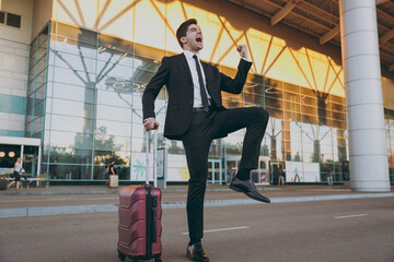 Wall Mural - Bottom view full length young traveler brunet businessman man in black dinner suit stand outside at international airport terminal with suitcase valise do winner gesture Air flight business concept.