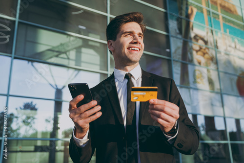 Bottom view traveler businessman man wear black suit stand outside at international airport terminal use mobile cell phone hold credit bank card doing online payment Air flight business trip concept