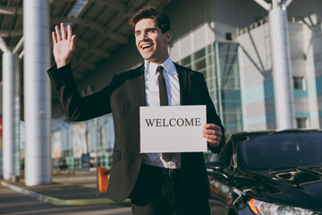 Wall Mural - Bottom view young cheerful friendly traveler businessman man 20s in black suit stand outside at international airport terminal hold card sign with welcome title text Air flight business trip concept