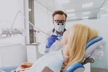 Young european man doctor in dentist binocular loupe mask drills tooth of patient woman with caries decay sitting at dentist office chair indoor cabinet near stomatologist Healthcare enamel treatment