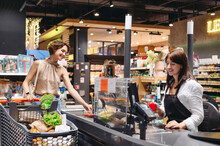 Young satisfied happy woman 20s wear casual clothes shopping at supermaket with grocery cart stand at store checkout pays for groceries cashier inside hypermarket Purchasing gastronomy food concept