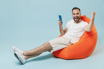 Wall Mural - Full length young happy man in white t-shirt sit in bag chair using mobile cell phone do winner gesture clench fist rest relax isolated on plain pastel light blue color background studio portrait.