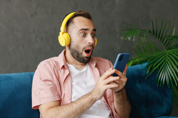 Canvas Print - Young shocked surprised amazed confused man in casual clothes headphones listen to music use hold mobile cell phone read news sit on blue sofa at home flat indoors rest relax on weekends free time