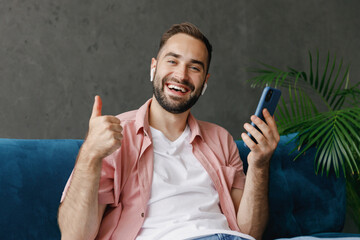 Wall Mural - Young smiling happy fun calm man in casual clothes earphones listen to music hold in hand use mobile cell phone show thumb up sitting on blue sofa at home flat indoor rest relax on weekends free time.