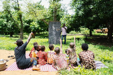 Mixed group of black children sitting outdoors, eagerly volunteering to answer their teacher's questions during class in an African community school