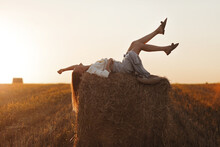 Young Woman With Long Hair, Wearing Jeans Skirt, Light Shirt Is Lying On Straw Bale In Field In Summer On Sunset. Female Portrait In Natural Rural Scene. Environmental Eco Tourism Concept.