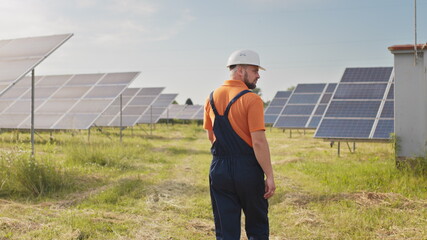 Wall Mural - Maintenance assistance technical worker in uniform is checking an operation and efficiency performance of photovoltaic solar panels. Construction engineer walks between solar panels on field station.