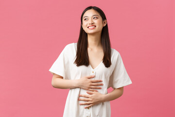 Beauty, people emotions and summer leisure and vacation concept. Pleased smiling asian girl looking dreamy as sky and rubbing belly, got rid of cramps or stomach ache with medicine, pink background