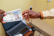 An African hand receiving Nigerian Naira notes, cash or currency with phone on a laptop in the background