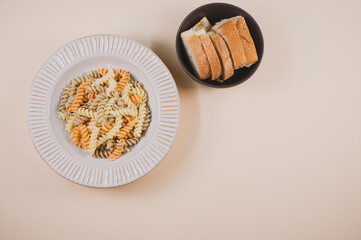 Wall Mural - Plate of pasta twists and a bowl of sliced bread on a beige background with free space for text