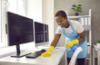 © Studio Romantic - Cheerful smiling cleaner carefully wipes the dust from the table in a bright office. African American woman in rubber gloves, apron and with a spray in her hands cleans the desktop with computers.