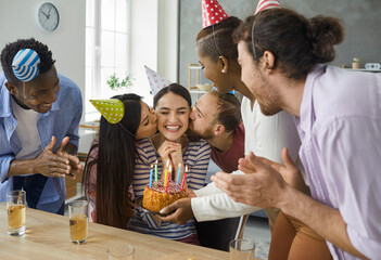 Multiethnic group of cheerful friends in cone caps kissing happy birthday girl at her birthday party at home. Young woman gets congratulations, kisses, presents and sweet cake for her birthday