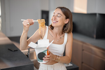 Young woman eating noodles with chopsticks from cardboard packaging in the kitchen at home. Takeaway asian food, ordering online concept