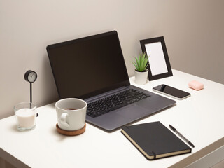Picture of laptop on a white table with a mug a clock a small bush a notebook a phone a picture frame and a headphone case